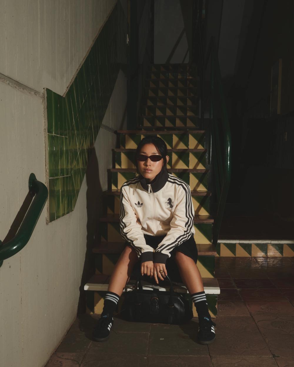 Modelo con chaqueta Adidas blanca y gafas de sol sentada en unas escaleras con azulejos