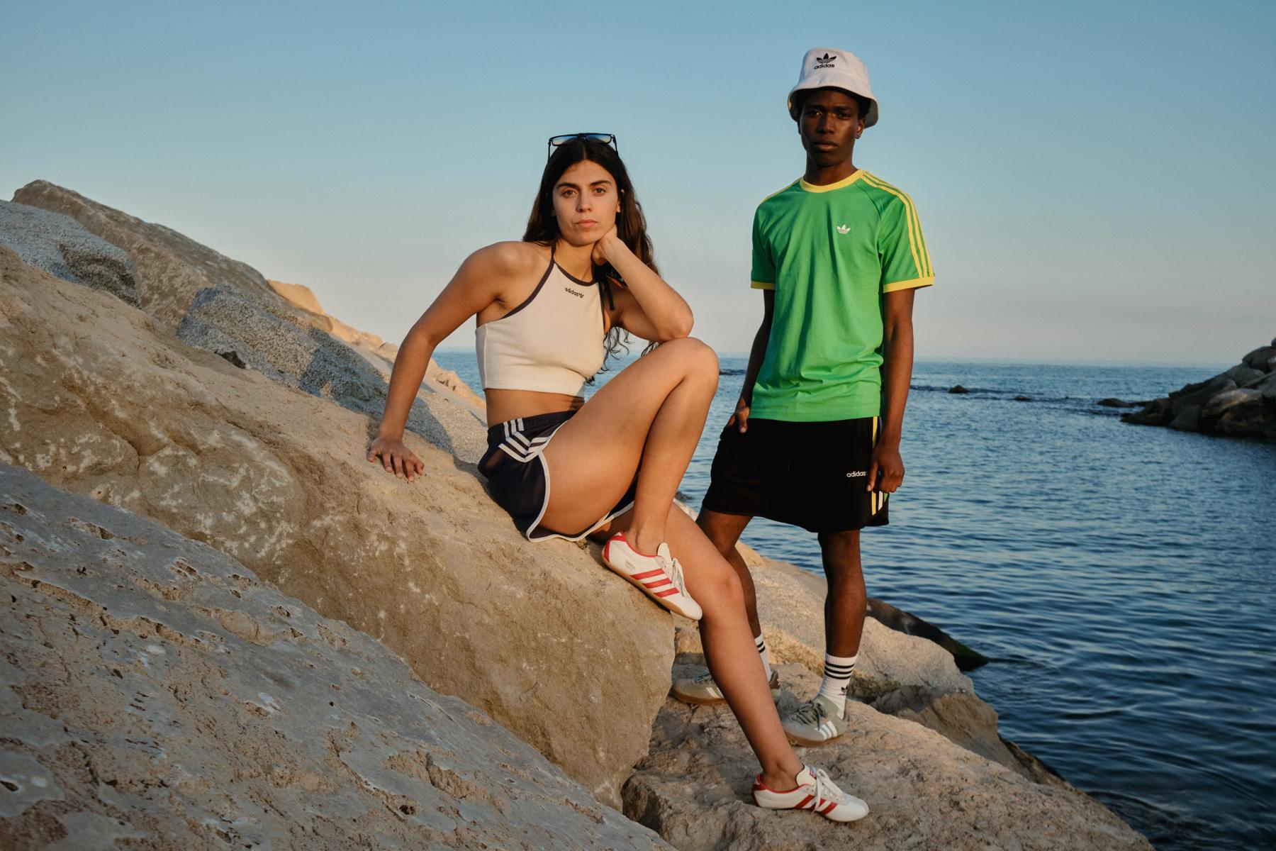 Dos modelos con ropa Adidas posan sobre unas rocas junto al mar al atardecer