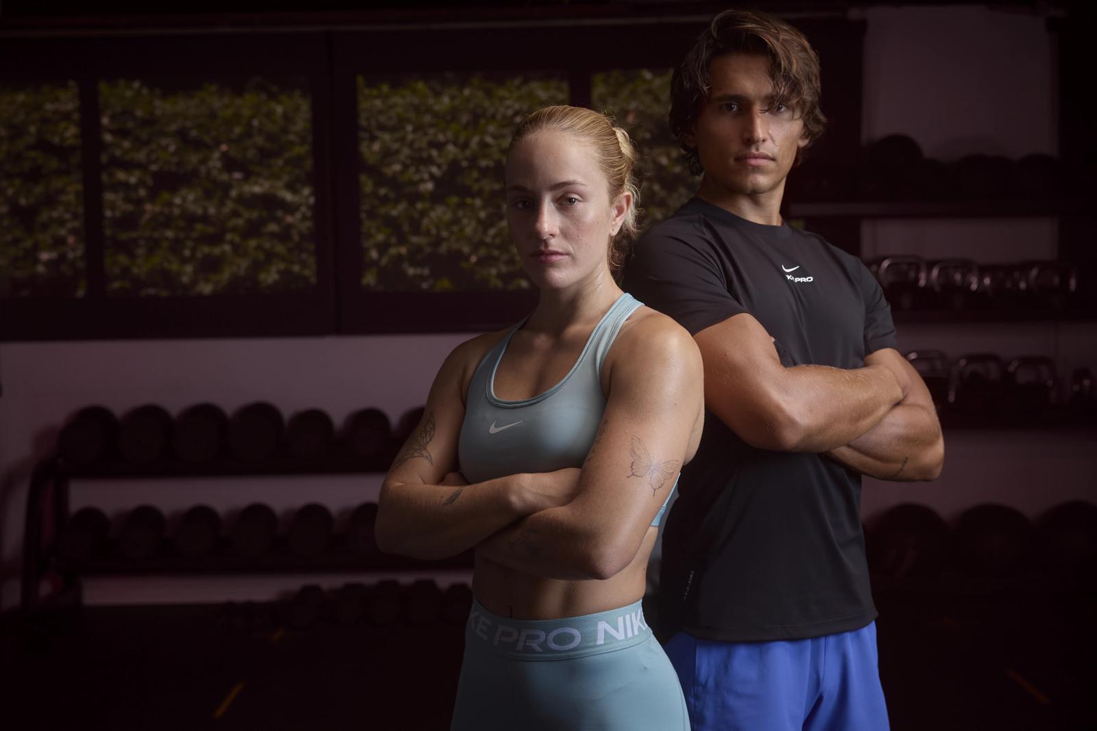 Una chica y un chico con ropa deportiva posan espalda con espalda y brazos cruzados en un gimnasio